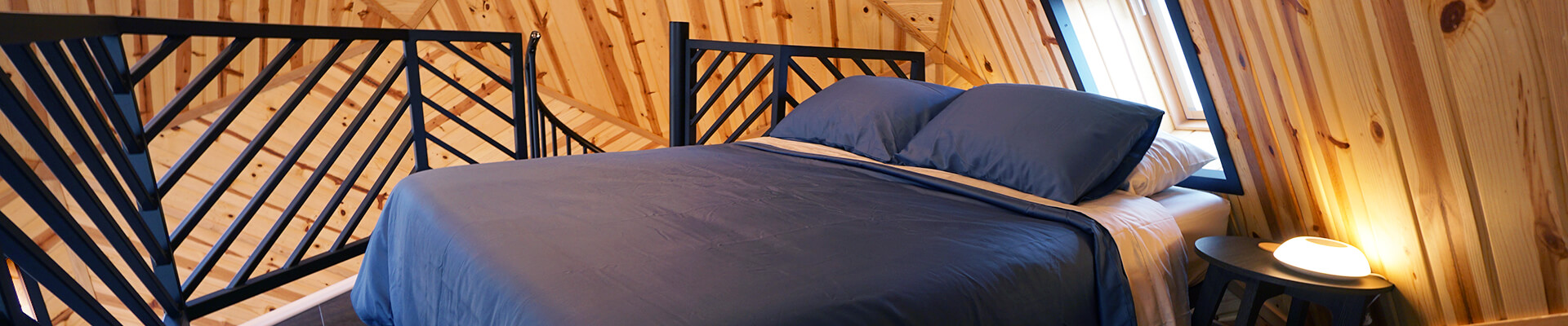 A close-up view of the interior geodesic dome loft, showcasing a queen bed next to a night table with a glowing lamp. The cozy space emphasizes a warm and inviting atmosphere.