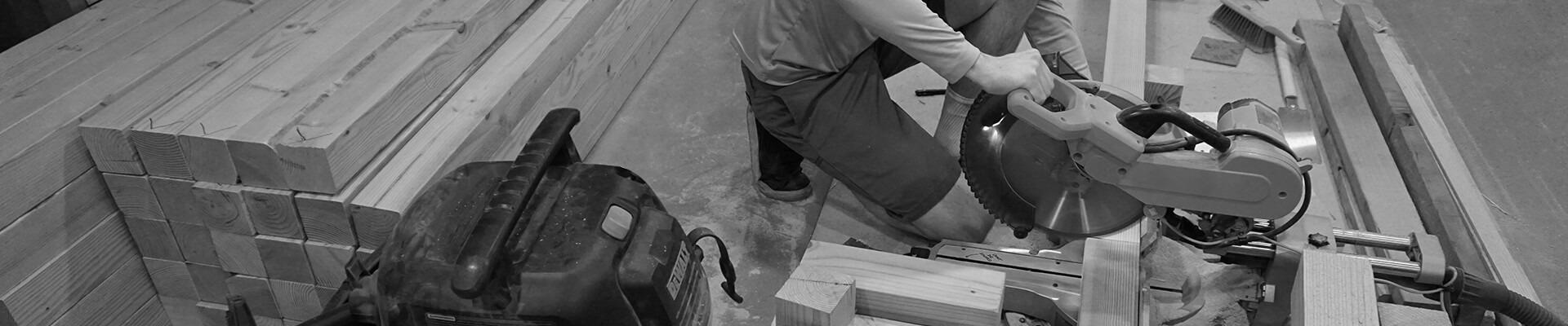 A black and white photo of the owner cutting wood on the floor of a garage, with material stacked up behind him.