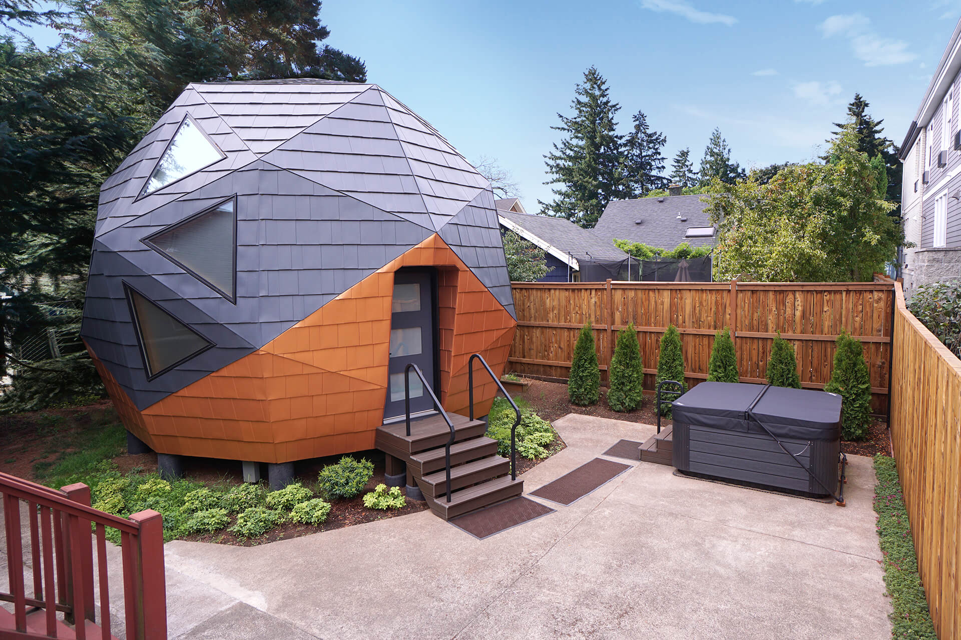 Wide shot of the mini geodesic dome home featuring a hot tub, privacy trees, and a cedar fence.