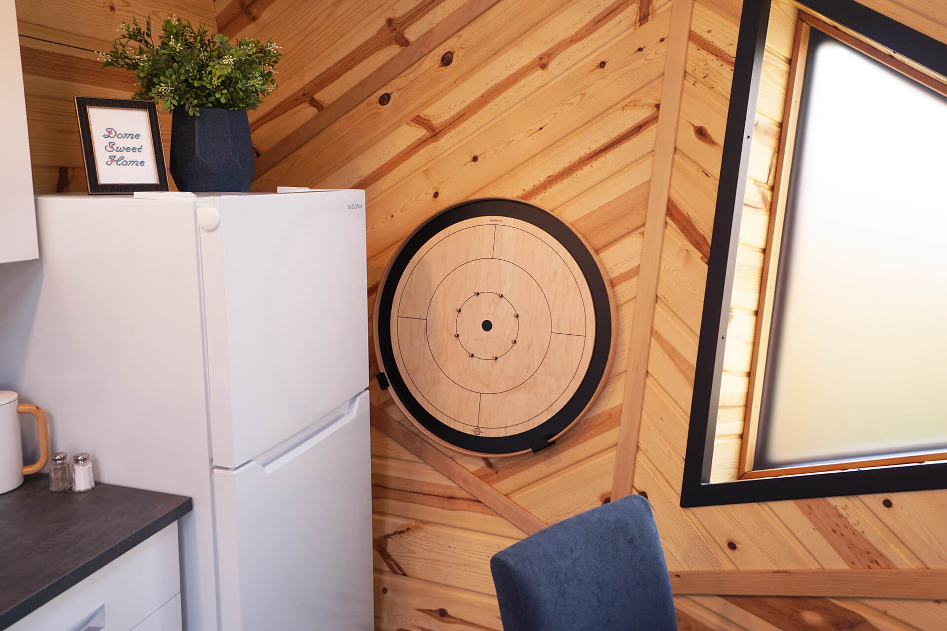 Crokinole board game mounted on the wall near the fridge, which is topped with a plant and an embroidery reading 'Dome Sweet Home'.