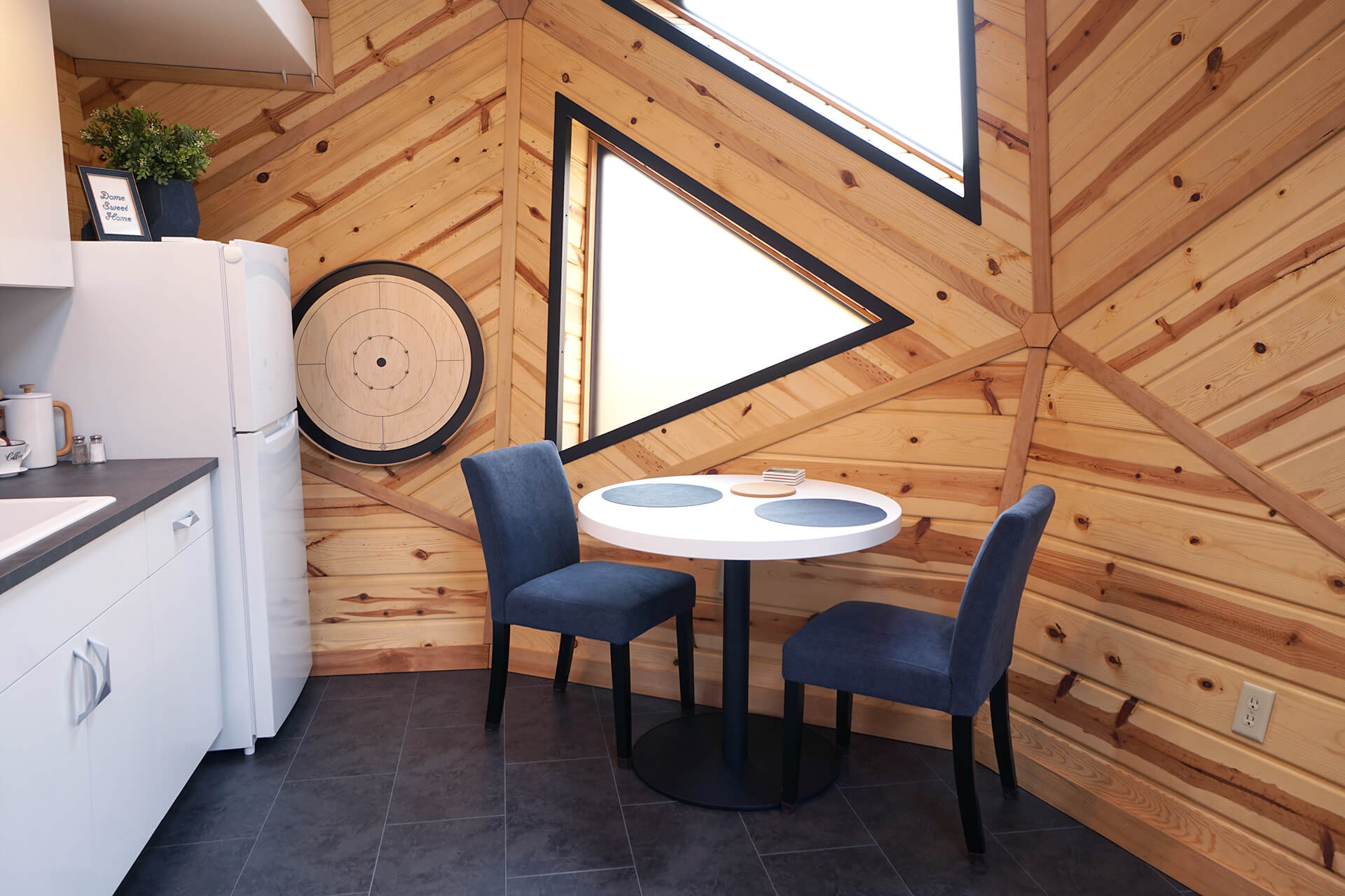 Dining area featuring a white round table in front of a large frosted triangular window, two blue chairs, a refrigerator, and a crokinole board game on the knotty pine wall.