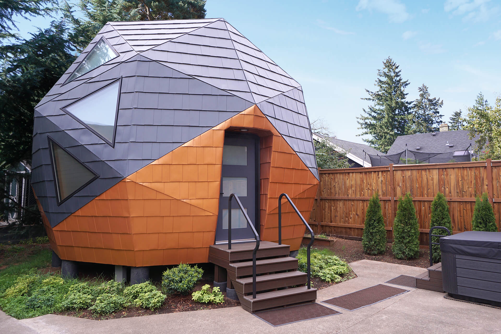 A mini geodesic dome home in a backyard, showcasing a modern and eco-friendly design with a four step front porch, surrounded by greenery and concrete.