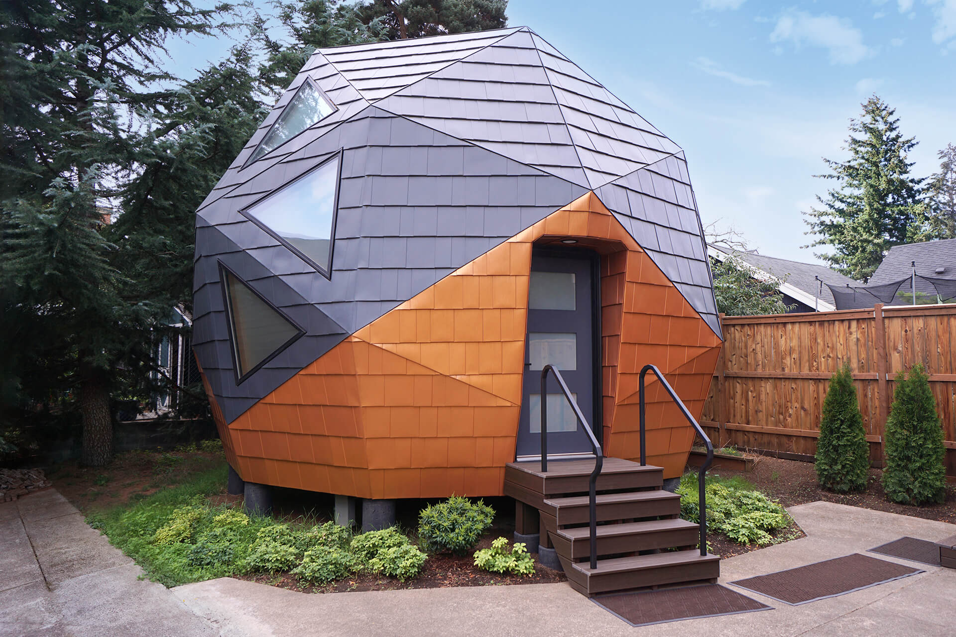 Mini geodesic dome home by Dodeca Domes, featuring vibrant orange metal siding and a dark grey shingle roof, with a unique porch adorned with angular handrails, all surrounded by lush greenery and concrete pathways.