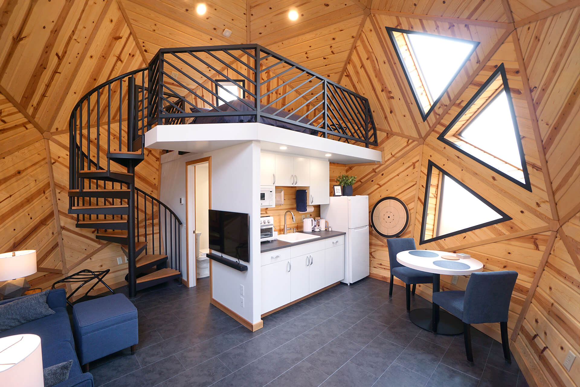 Wide view of the interior showcasing the sleeping loft, kitchenette, dining area, couch, bathroom, spiral staircase, slate flooring, and knotty pine walls.