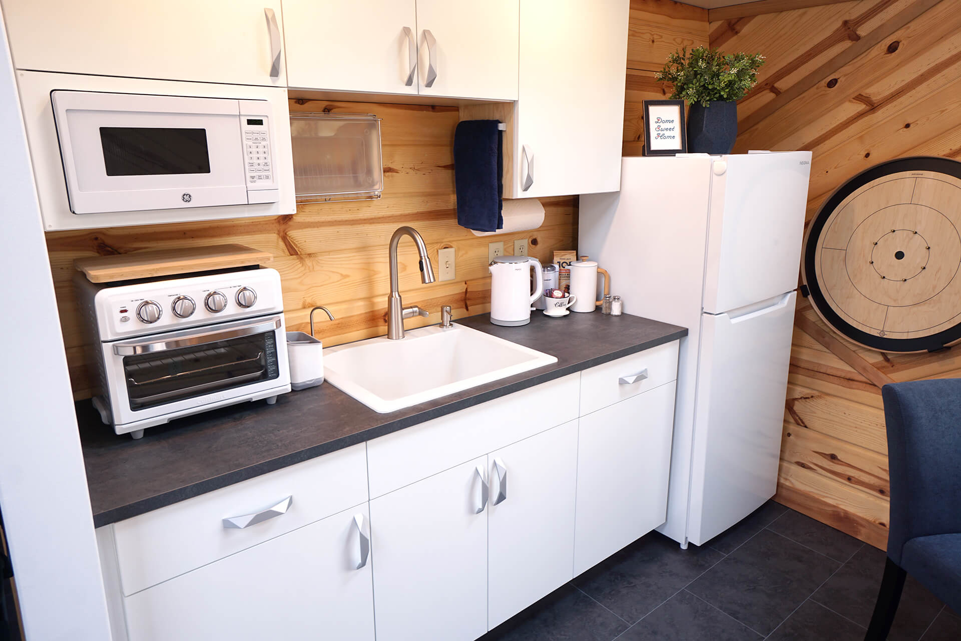 Kitchenette featuring white cabinets, a tabletop oven, built-in microwave, electric kettle, dish drying rack, and a white sink on a dark countertop that matches the composite slate flooring.