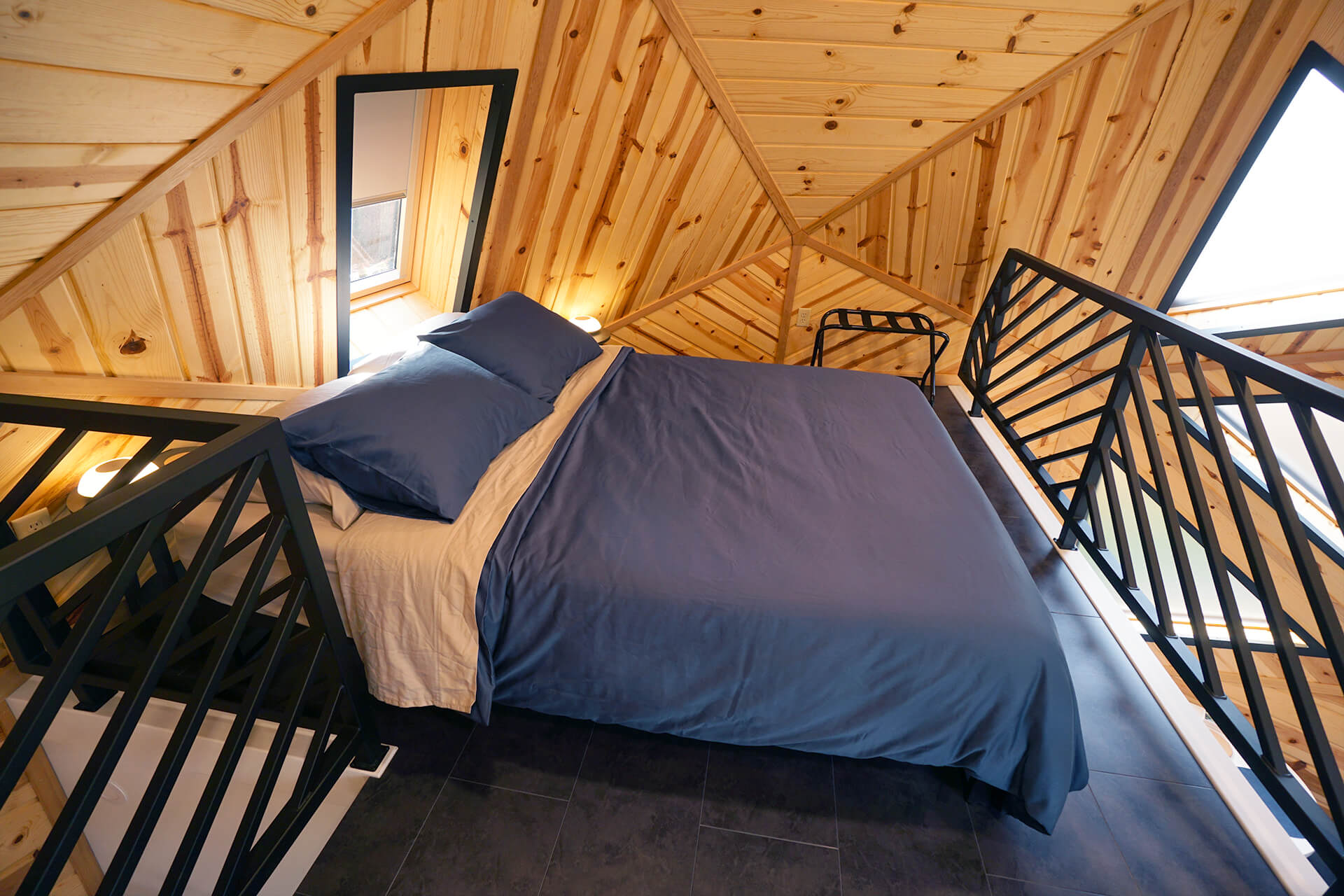 View of the sleeping loft, featuring a queen-sized bed with blue linens, a folding luggage rack, and side tables adorned with glowing dome-shaped lamps.